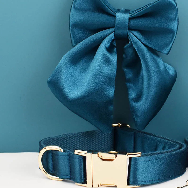 Collier bleu pétrole pour chien avec grand nœud détachable et anneau métal doré présenté devant un fond bleu et blanc.