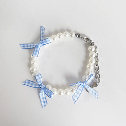 Collier fantaisie pour chiot composé de perles blanches et rubans à carreaux bleu et blanc avec chaînette de réglage et mousquetou en métal sur fond clair.