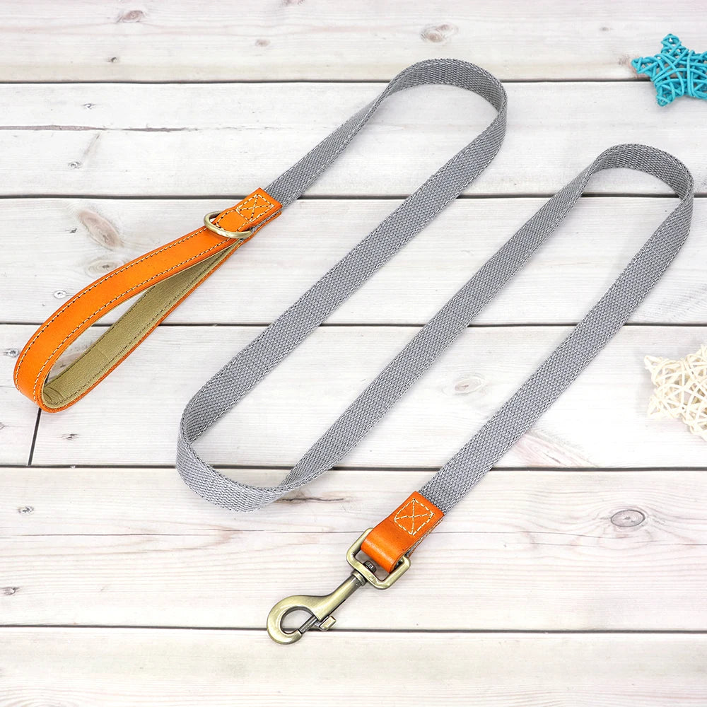 Laisse grise pour chien avec poignée en cuir orange et mousqueton métal présenté sur un plancher en bois gris dans le fond.