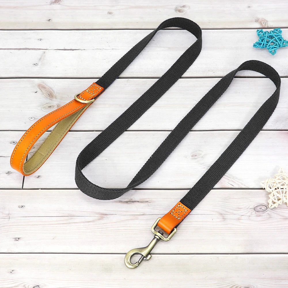 Laisse noire pour chien avec poignée en cuir orange et mousqueton métal présenté sur un plancher en bois gris dans le fond.